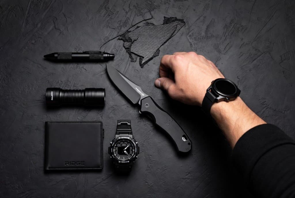 Flat lay of black EDC gear — knife, flashlight, wallet, pen, and watch on a dark textured surface